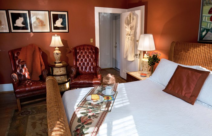 A bedroom at Bischwind Villa featuring a bed with a breakfast tray, two leather wingback chairs, and three cat portraits on a red wall.