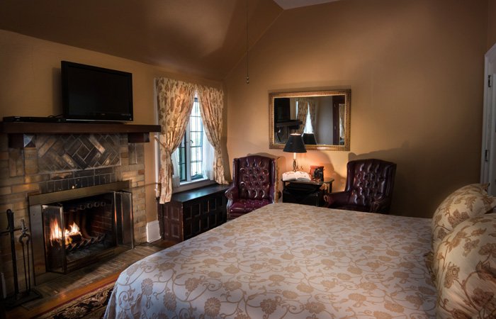 A guest room at Bischwind Villa featuring a lit fireplace, two leather armchairs, and a large bed with a patterned coverlet.