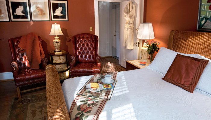 A hotel bedroom featuring Bischwind Villa policies with two leather armchairs, a white bed with a breakfast tray, and a bathrobe hanging on the door.