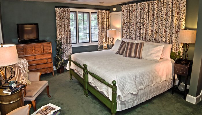 A bedroom with a green bed frame, a white quilt, patterned floor-to-ceiling curtains, a wooden dresser with a television, and two bedside tables with lamps, reflecting the style found in Bischwind villa policies.