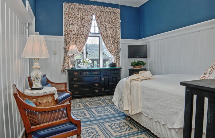 Bedroom with blue walls and white wainscoting at Bischwind Villa, featuring a bed with a white quilt, two wooden armchairs, and a dark wood dresser beneath a window.