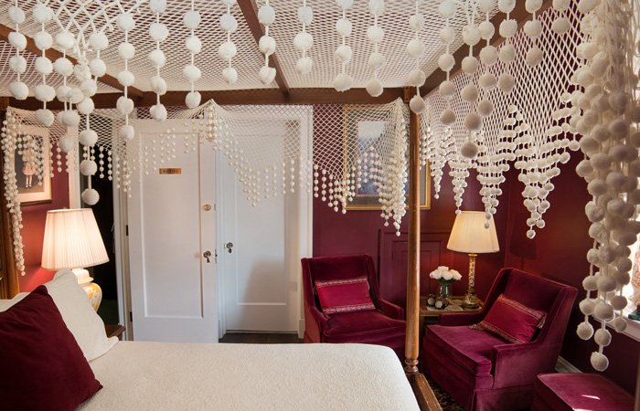 A four-poster bed with a decorative white pom-pom canopy in a Bischwind Villa guest room, featuring deep red walls and two matching velvet armchairs.