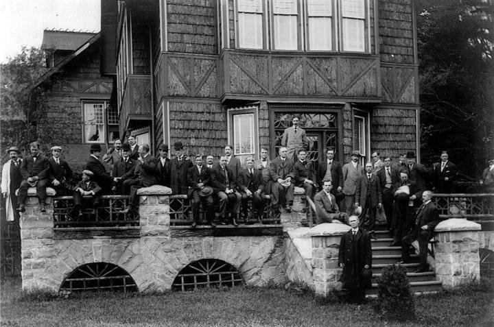 A large group of men in formal suits posing for a photograph in front of the Bischwind Villa, a multi-story house with a stone foundation.