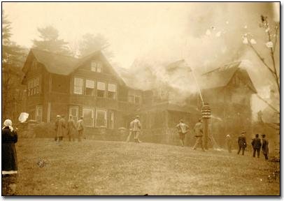 A sepia-toned historical photograph showing a group of people standing on a lawn watching the Bischwind villa engulfed in smoke.