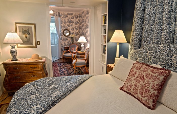 A bedroom at Bischwind Villa featuring a bed with blue and white patterned bedding and a red toile pillow, leading to a smaller sitting area with two chairs and blue and white toile wallpaper.