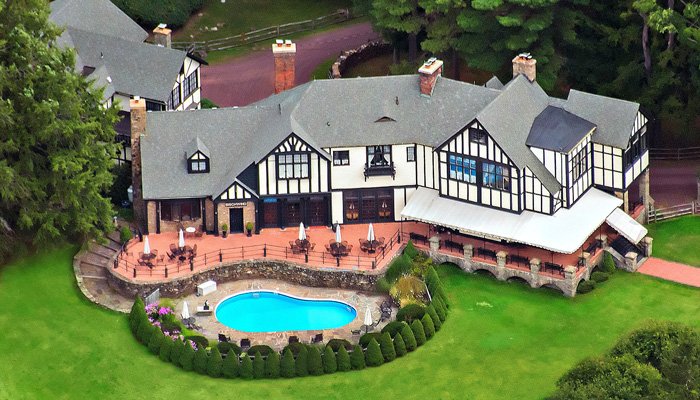Aerial view of a large Tudor-style estate with a curved patio, an outdoor swimming pool, and sprawling green lawns.