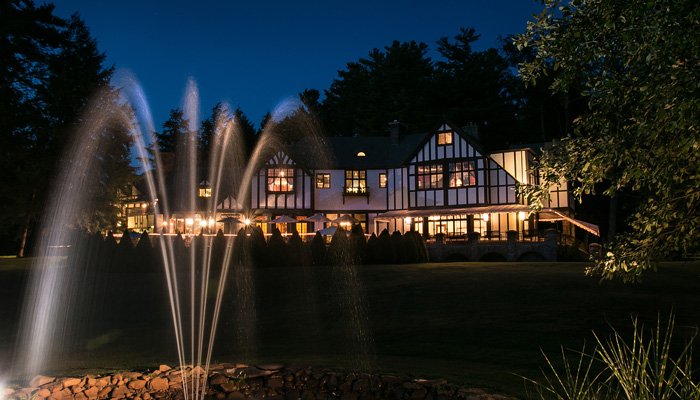 A large Tudor-style mansion illuminated at twilight, viewed across a lawn with a fountain in the foreground.